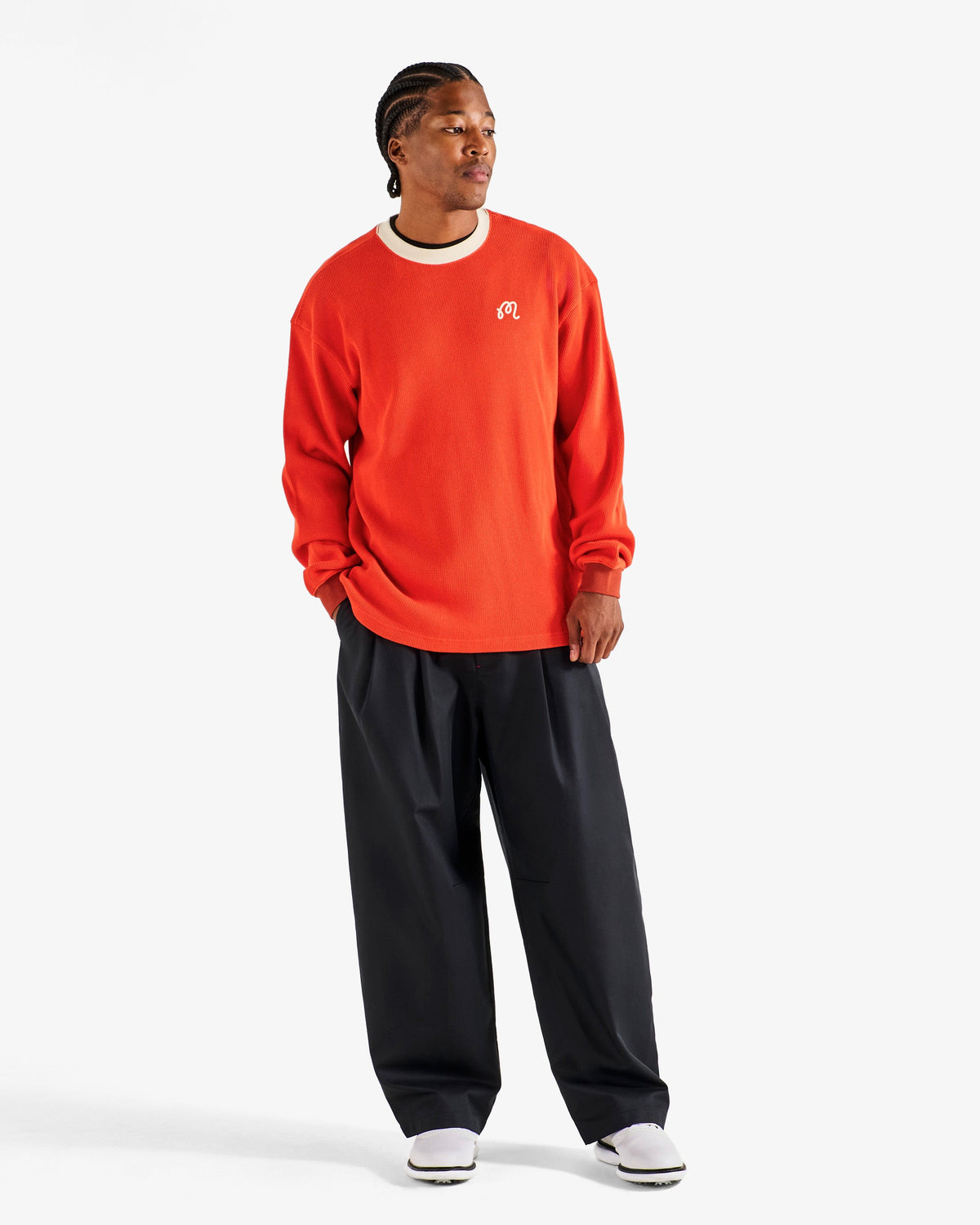 Model wearing the Malbon Golf Gorse Heritage Long Sleeve in orange red with black trousers, front three-quarter view showing the relaxed dropped-shoulder fit
