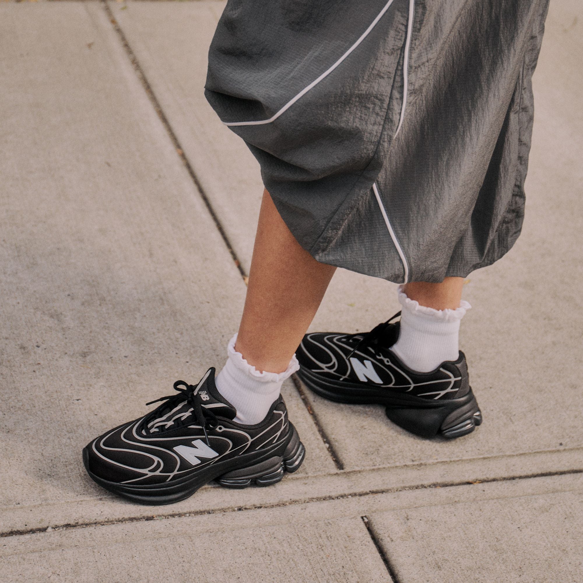 New Balance 5030 Black with Silver Metallic worn on foot on pavement with grey trousers